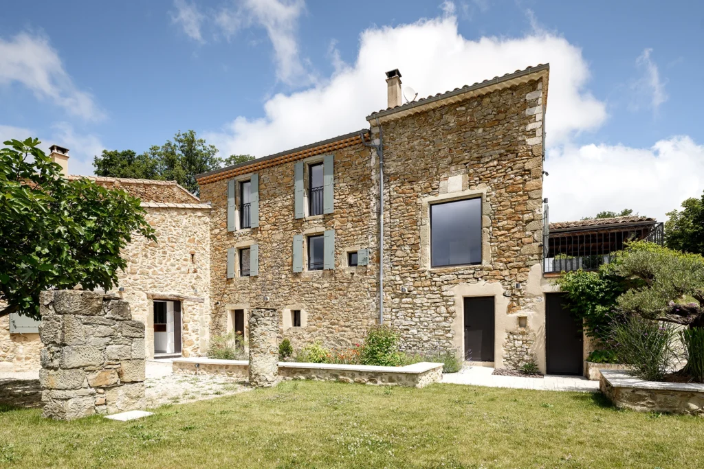 Rénovation complète d’une maison de famille – Drôme
Nichée au cœur des paysages de la Drôme, cette maison a fait l’objet d’une rénovation intégrale. Le projet a consisté à redonner vie à une bâtisse aux volumes autrefois fermés et cloisonnés, pour en révéler tout le potentiel architectural et spatial.
L’intervention a porté sur l’ensemble de l’enveloppe : les façades ont été restaurées, les ouvertures repensées et créées pour offrir de nouveaux cadrages sur le paysage, mettant en valeur la pierre existante et la richesse du contexte environnant.
À l’intérieur, les espaces ont été entièrement réorganisés. Le plan initial, fragmenté et contraint, a laissé place à des enchaînements fluides, où les pièces dialoguent entre elles et s’ouvrent généreusement sur l’extérieur. Chaque ouverture devient un tableau vivant, cadrant une portion de nature : champs de blé, forêt, lumière changeante.
Le cœur de la maison se structure autour de volumes entremêlés. Une cheminée centrale, élément à la fois architectural et domestique, relie les grands volumes de la cuisine en double hauteur et le salon, plus intime et chaleureux. Dans une autre aile, à l’étage, une seconde cuisine a été aménagée. Elle prolonge l’esprit du projet : fonctionnalité discrète, ouverture maîtrisée, cadrages sensibles sur le paysage.
Les ambiances varient selon les espaces : une chambre se déploie autour d’un vide intérieur, jouant avec les hauteurs et les couleurs ; le salon TV, équipé d’une bibliothèque et de rangements intégrés, offre un lieu de retrait, propice au calme tout en restant connecté à l’extérieur.
Le choix des matériaux renforce l’identité sobre et chaleureuse du lieu : pierre naturelle, travertin, parquet en chêne et surfaces en solid surface s’équilibrent pour créer une atmosphère douce, intemporelle et raffinée.
Les ouvertures ont été dessinées pour favoriser la lumière naturelle et créer des liens visuels avec le paysage, tout en accompagnant les usages quotidiens. À l’extérieur, une piscine s’insère discrètement dans le jardin paysager, entourée de terrasses orientées pour profiter du soleil tout au long de la journée. La maison se fond progressivement dans son environnement, entre les cultures et les bois, dans un dialogue apaisé avec le site.
Enfin, les espaces de nuit, regroupés dans une aile plus en retrait, assurent calme et sérénité. Ils sont complétés par une salle de sport et un second salon dédié aux plus jeunes, pour que chacun puisse vivre la maison à son rythme, en toute liberté.