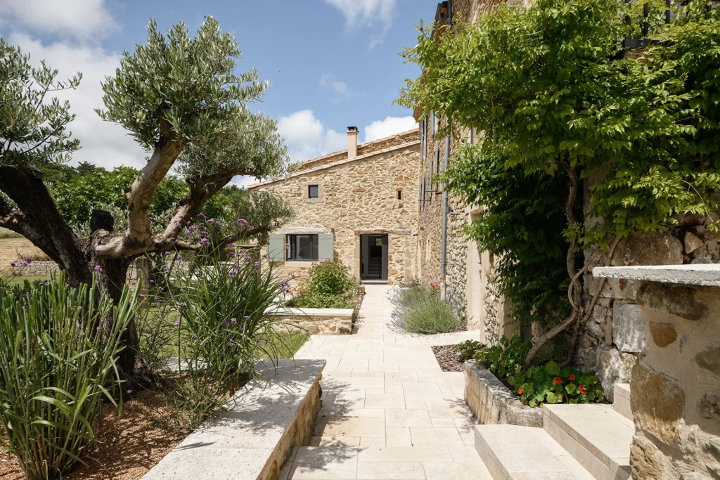 Rénovation complète d’une maison de famille – Drôme
Nichée au cœur des paysages de la Drôme, cette maison a fait l’objet d’une rénovation intégrale. Le projet a consisté à redonner vie à une bâtisse aux volumes autrefois fermés et cloisonnés, pour en révéler tout le potentiel architectural et spatial.
L’intervention a porté sur l’ensemble de l’enveloppe : les façades ont été restaurées, les ouvertures repensées et créées pour offrir de nouveaux cadrages sur le paysage, mettant en valeur la pierre existante et la richesse du contexte environnant.
À l’intérieur, les espaces ont été entièrement réorganisés. Le plan initial, fragmenté et contraint, a laissé place à des enchaînements fluides, où les pièces dialoguent entre elles et s’ouvrent généreusement sur l’extérieur. Chaque ouverture devient un tableau vivant, cadrant une portion de nature : champs de blé, forêt, lumière changeante.
Le cœur de la maison se structure autour de volumes entremêlés. Une cheminée centrale, élément à la fois architectural et domestique, relie les grands volumes de la cuisine en double hauteur et le salon, plus intime et chaleureux. Dans une autre aile, à l’étage, une seconde cuisine a été aménagée. Elle prolonge l’esprit du projet : fonctionnalité discrète, ouverture maîtrisée, cadrages sensibles sur le paysage.
Les ambiances varient selon les espaces : une chambre se déploie autour d’un vide intérieur, jouant avec les hauteurs et les couleurs ; le salon TV, équipé d’une bibliothèque et de rangements intégrés, offre un lieu de retrait, propice au calme tout en restant connecté à l’extérieur.
Le choix des matériaux renforce l’identité sobre et chaleureuse du lieu : pierre naturelle, travertin, parquet en chêne et surfaces en solid surface s’équilibrent pour créer une atmosphère douce, intemporelle et raffinée.
Les ouvertures ont été dessinées pour favoriser la lumière naturelle et créer des liens visuels avec le paysage, tout en accompagnant les usages quotidiens. À l’extérieur, une piscine s’insère discrètement dans le jardin paysager, entourée de terrasses orientées pour profiter du soleil tout au long de la journée. La maison se fond progressivement dans son environnement, entre les cultures et les bois, dans un dialogue apaisé avec le site.
Enfin, les espaces de nuit, regroupés dans une aile plus en retrait, assurent calme et sérénité. Ils sont complétés par une salle de sport et un second salon dédié aux plus jeunes, pour que chacun puisse vivre la maison à son rythme, en toute liberté.