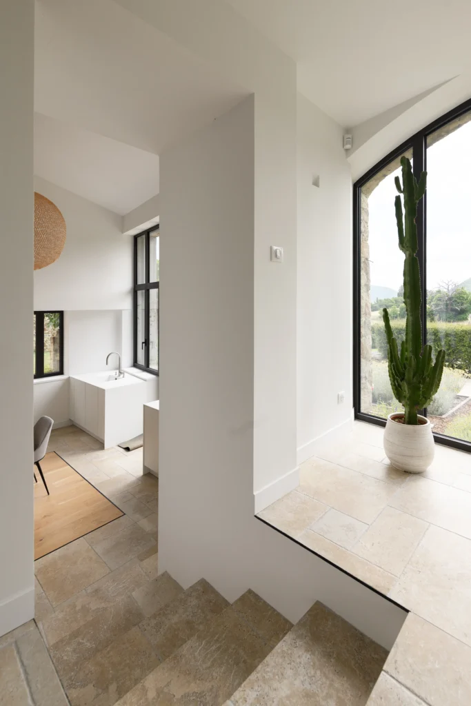 Rénovation complète d’une maison de famille – Drôme
Nichée au cœur des paysages de la Drôme, cette maison a fait l’objet d’une rénovation intégrale. Le projet a consisté à redonner vie à une bâtisse aux volumes autrefois fermés et cloisonnés, pour en révéler tout le potentiel architectural et spatial.
L’intervention a porté sur l’ensemble de l’enveloppe : les façades ont été restaurées, les ouvertures repensées et créées pour offrir de nouveaux cadrages sur le paysage, mettant en valeur la pierre existante et la richesse du contexte environnant.
À l’intérieur, les espaces ont été entièrement réorganisés. Le plan initial, fragmenté et contraint, a laissé place à des enchaînements fluides, où les pièces dialoguent entre elles et s’ouvrent généreusement sur l’extérieur. Chaque ouverture devient un tableau vivant, cadrant une portion de nature : champs de blé, forêt, lumière changeante.
Le cœur de la maison se structure autour de volumes entremêlés. Une cheminée centrale, élément à la fois architectural et domestique, relie les grands volumes de la cuisine en double hauteur et le salon, plus intime et chaleureux. Dans une autre aile, à l’étage, une seconde cuisine a été aménagée. Elle prolonge l’esprit du projet : fonctionnalité discrète, ouverture maîtrisée, cadrages sensibles sur le paysage.
Les ambiances varient selon les espaces : une chambre se déploie autour d’un vide intérieur, jouant avec les hauteurs et les couleurs ; le salon TV, équipé d’une bibliothèque et de rangements intégrés, offre un lieu de retrait, propice au calme tout en restant connecté à l’extérieur.
Le choix des matériaux renforce l’identité sobre et chaleureuse du lieu : pierre naturelle, travertin, parquet en chêne et surfaces en solid surface s’équilibrent pour créer une atmosphère douce, intemporelle et raffinée.
Les ouvertures ont été dessinées pour favoriser la lumière naturelle et créer des liens visuels avec le paysage, tout en accompagnant les usages quotidiens. À l’extérieur, une piscine s’insère discrètement dans le jardin paysager, entourée de terrasses orientées pour profiter du soleil tout au long de la journée. La maison se fond progressivement dans son environnement, entre les cultures et les bois, dans un dialogue apaisé avec le site.
Enfin, les espaces de nuit, regroupés dans une aile plus en retrait, assurent calme et sérénité. Ils sont complétés par une salle de sport et un second salon dédié aux plus jeunes, pour que chacun puisse vivre la maison à son rythme, en toute liberté.